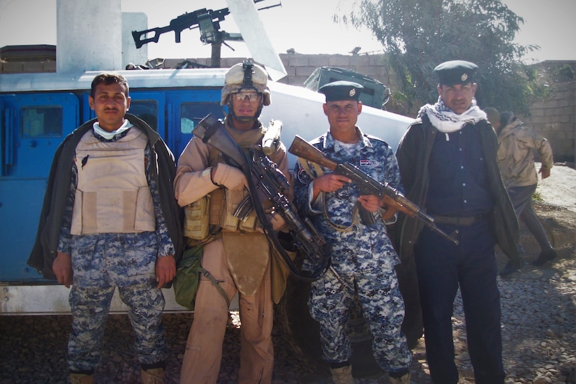 Four men dressed in different uniforms pose for a photo with an armored vehicle behind them. The two men in the middle are holding weapons.