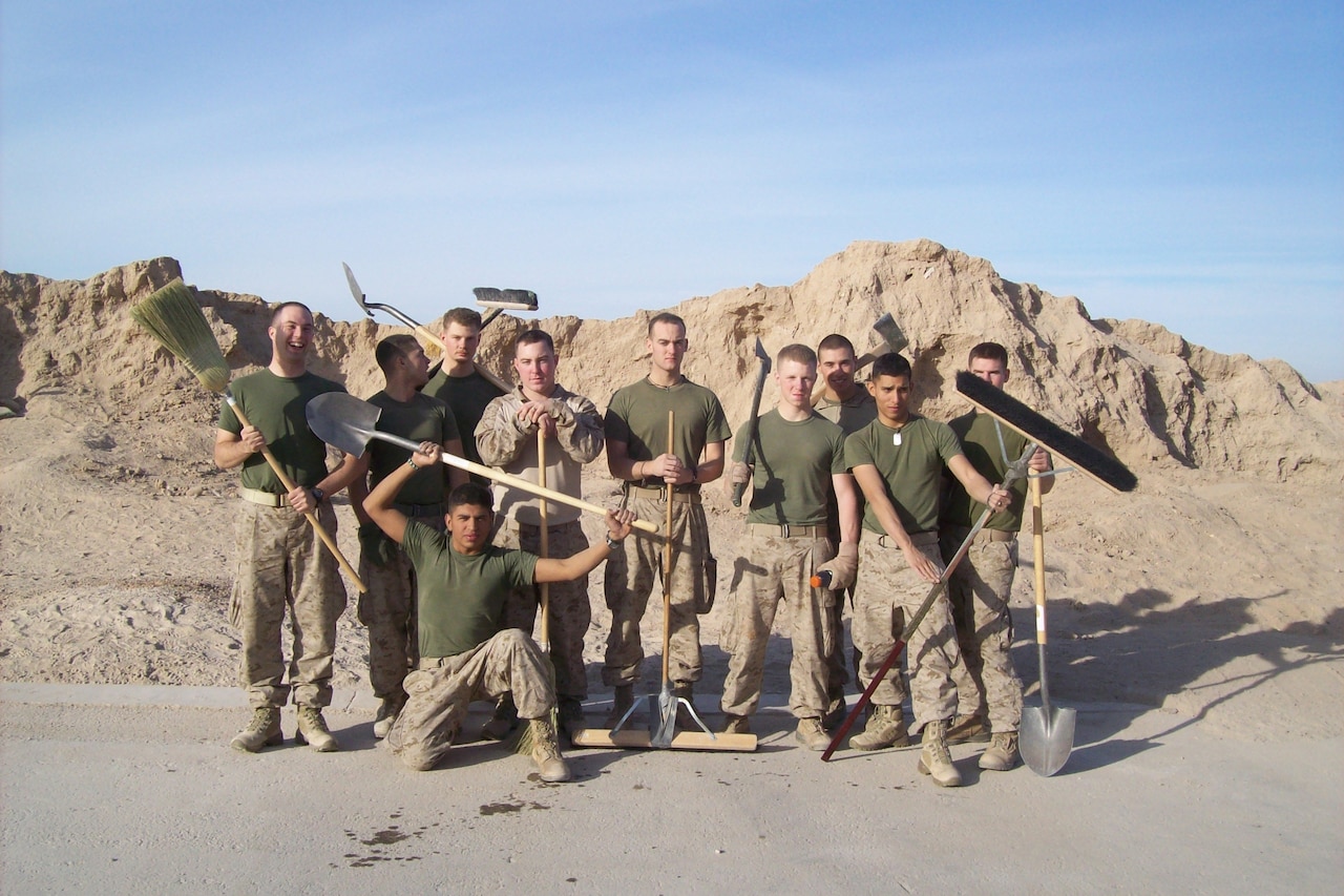 Ten Marines holding cleaning and digging tools pose for a photograph with large mounds of sand in the background.
