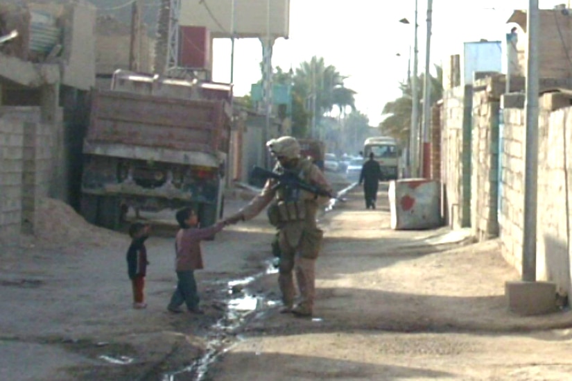 Walking down a narrow, dirt street lined with walls made of concrete blocks, a Marine in combat gear stops to shake the hand of a  small child while another child looks on.from a distance.