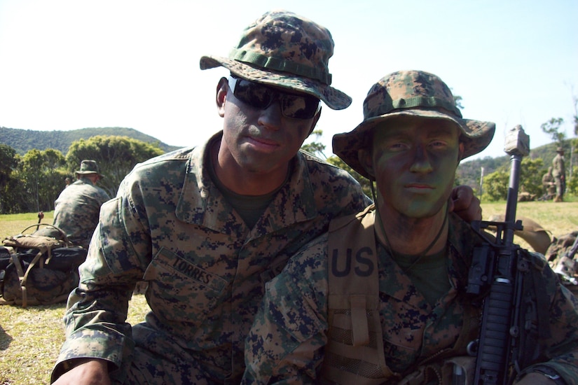 Two Marines,  wearing camouflage uniforms and camouflage face paint, pose for a photo in a field with trees in the background.
