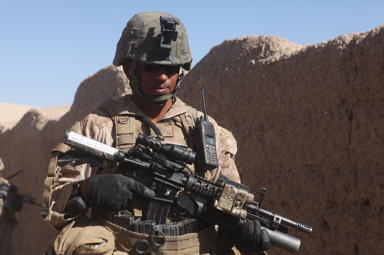 A Marine in full combat gear, helmet and rifle walks beside a desert wall.