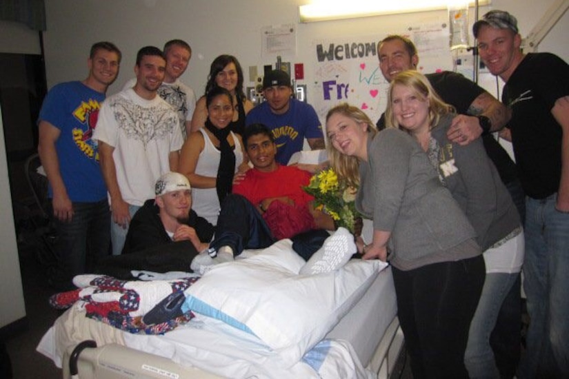 Eleven people surround a young man lying in a hospital bed with his leg bandaged and propped up on a pillow.