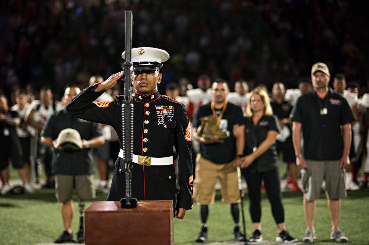 A Marine in dress uniform salutes a weapon pointing downward atop a wooden box on a football field. Four people, including a woman and two men with prosthetic legs look on from behind; football players stand in the background.