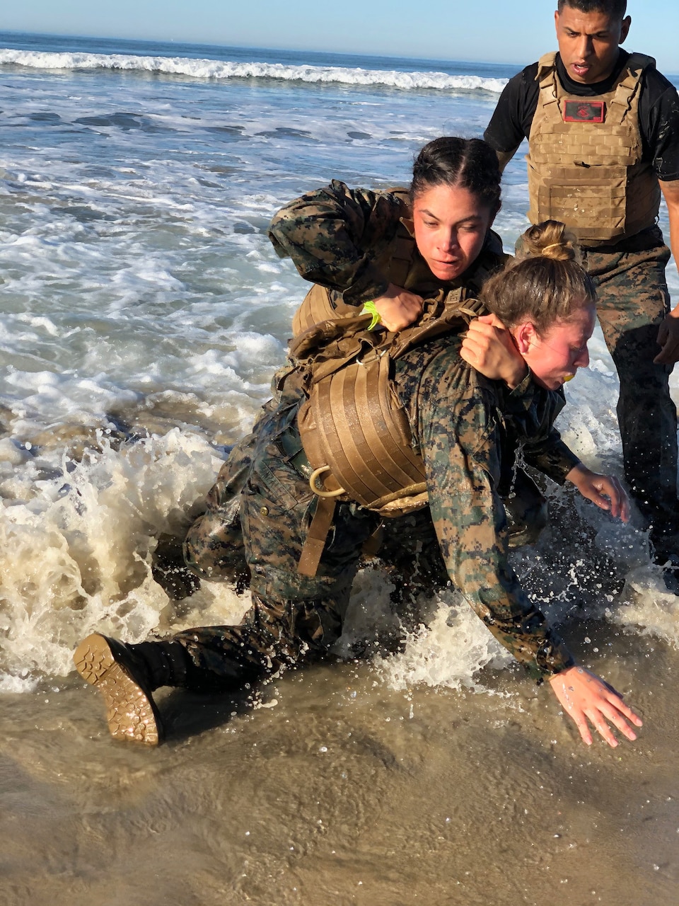 A male Marine watches two female Marines who are practicing martial arts in the ocean near a beach.