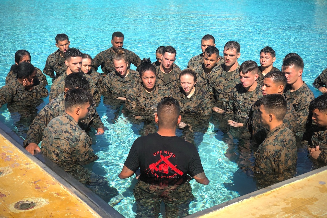 A Marine instructor with his back to the camera instructs about two dozen Marines in fatigues who are huddled in front of him in a swimming pool.