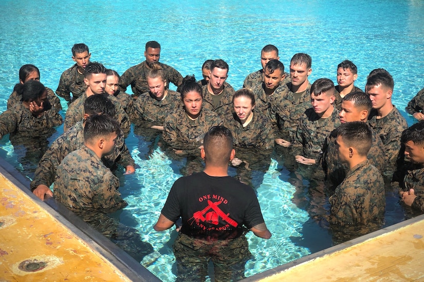 A Marine instructor with his back to the camera instructs about two dozen Marines in fatigues who are huddled in front of him in a swimming pool.