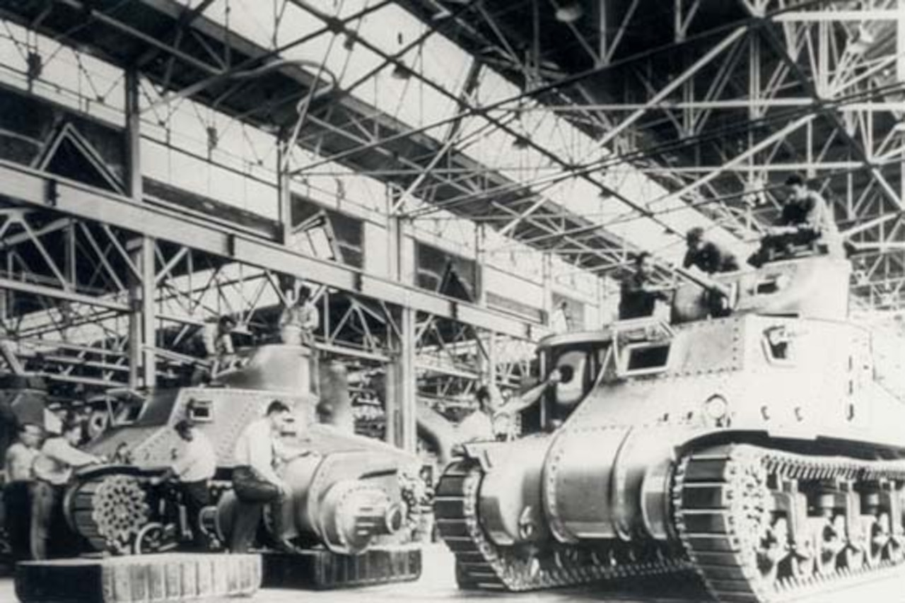 A black and white photos shows men constructing military tanks inside a large production facility.
