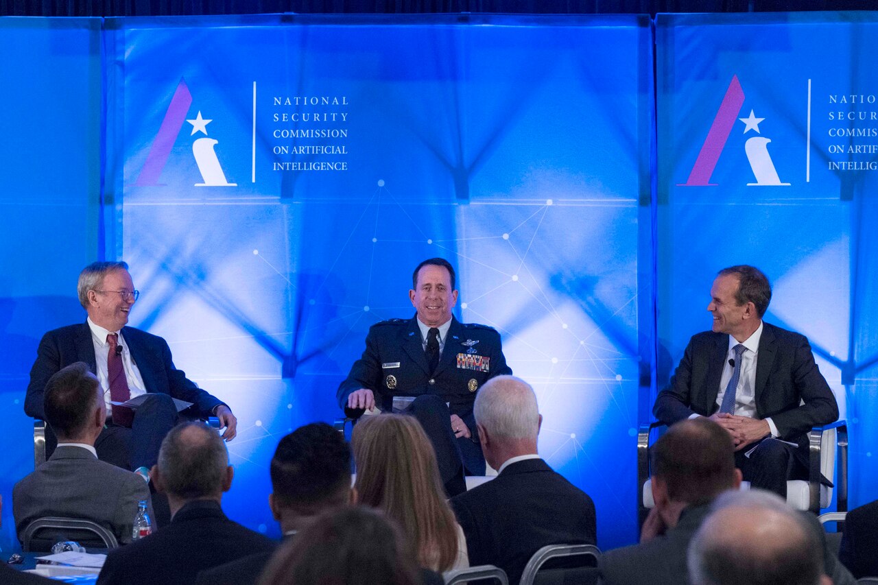 Three men sit in chairs on a stage in front of an audience.