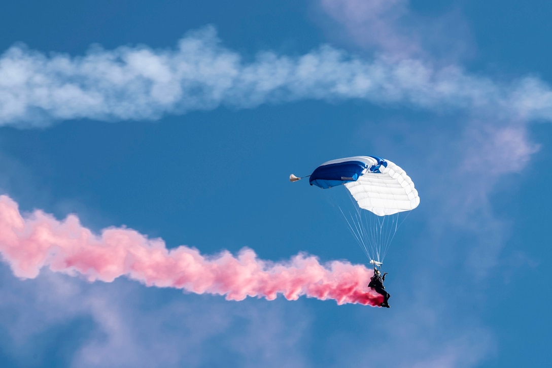 A service member glides through the sky with a parachute open as smoke blows from behind.