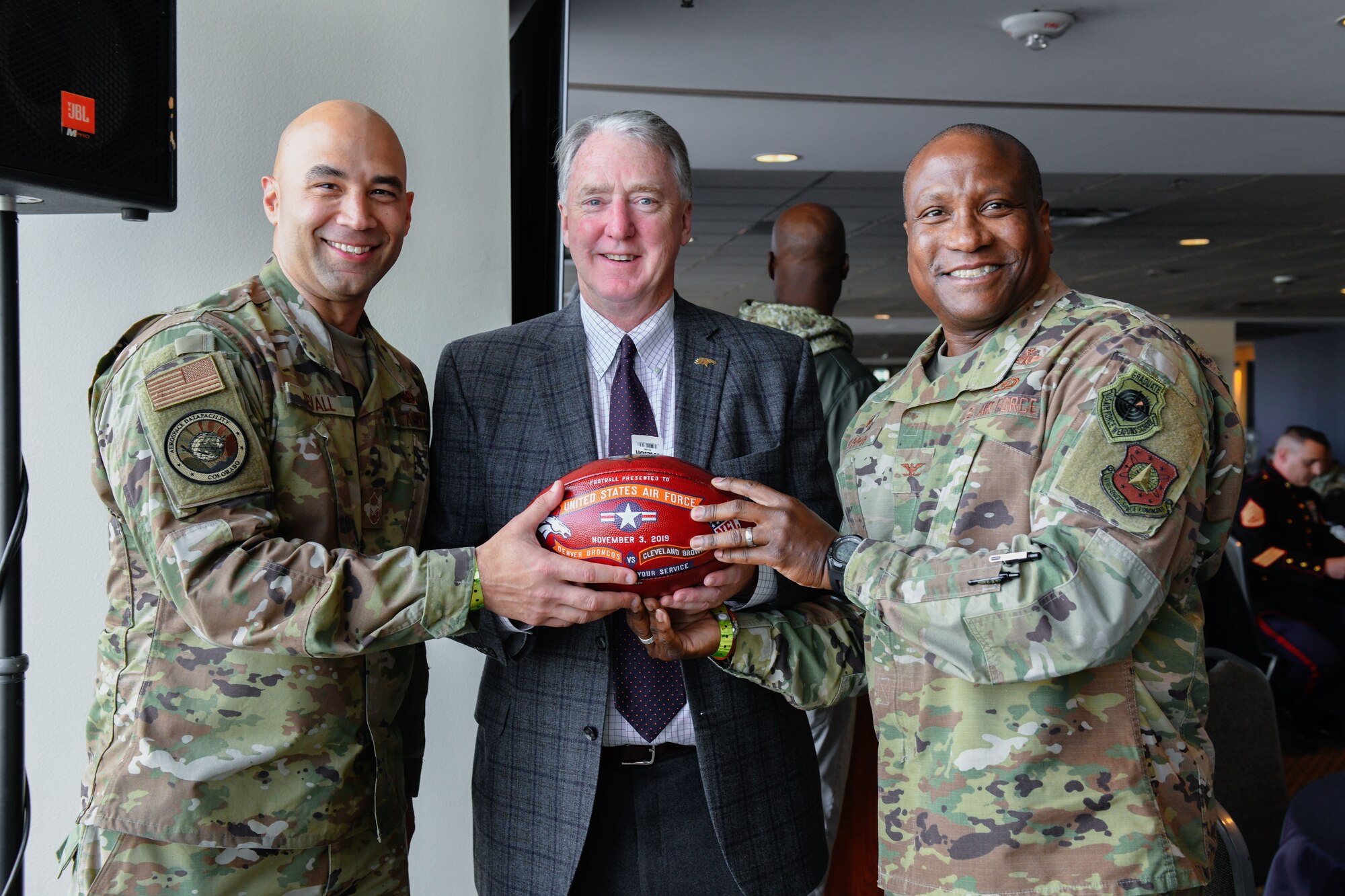 U.S. Air Force Chief Master Sgt. Robert Devall (left), the Aerospace Data Facility-Colorado senior enlisted leader, and Col. Devin Pepper (right), the 460th Space Wing commander, pose for a photo with Joe Ellis, the Denver Broncos president and chief executive officer, Nov. 3, 2019, at Empower Field at Mile High in Denver.