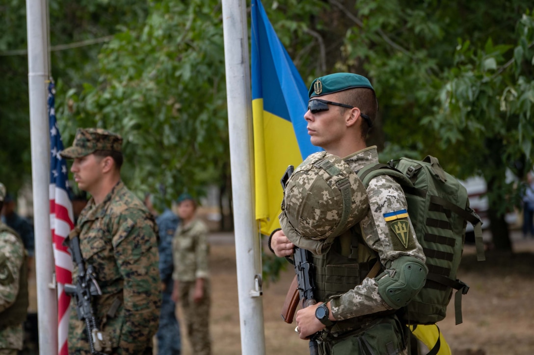 A service member in camouflage uniform stands next to the Ukranian flag.