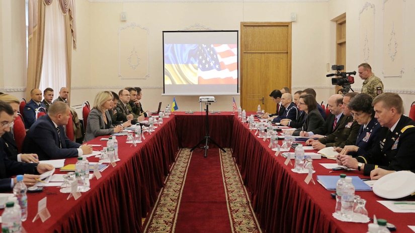 People in business suits and military uniforms sit at long tables facing each other.