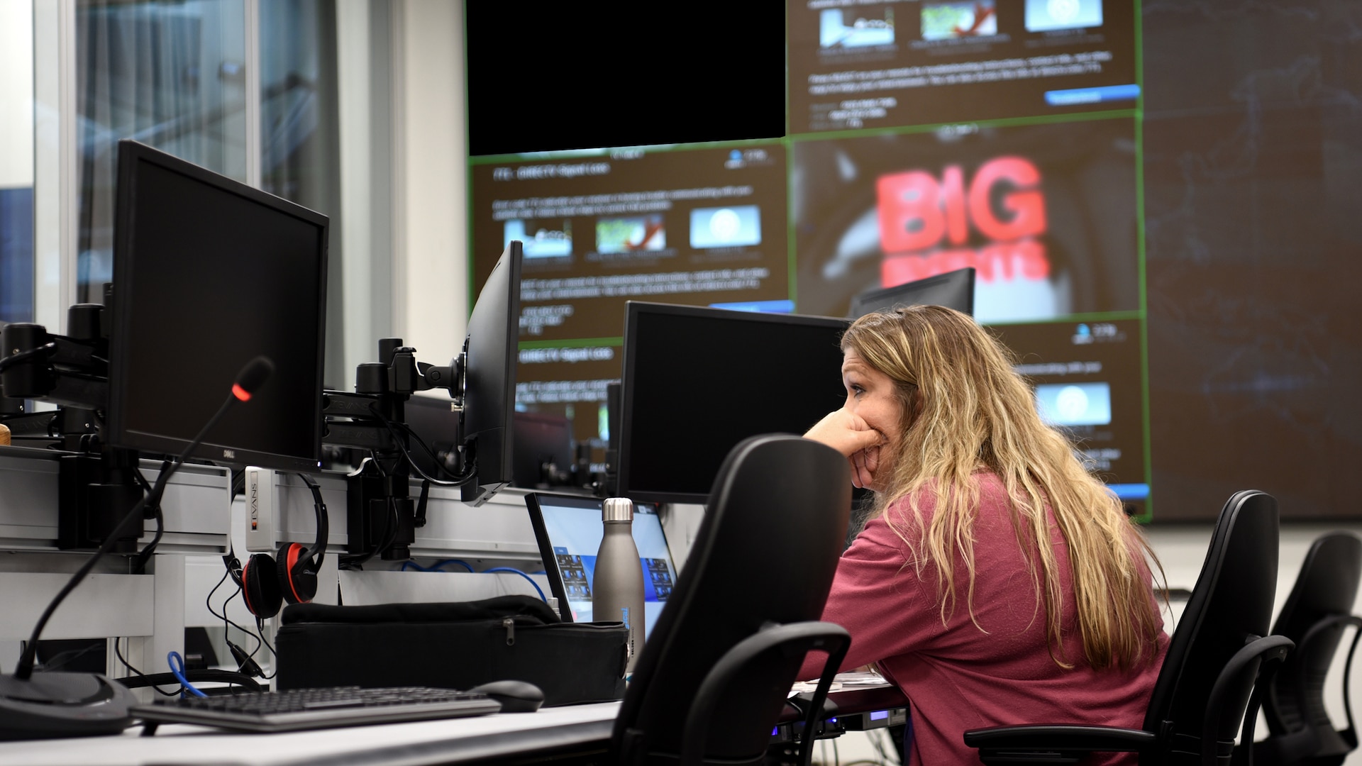 Woman sits in front of a computer screen with large TV monitors in the background.