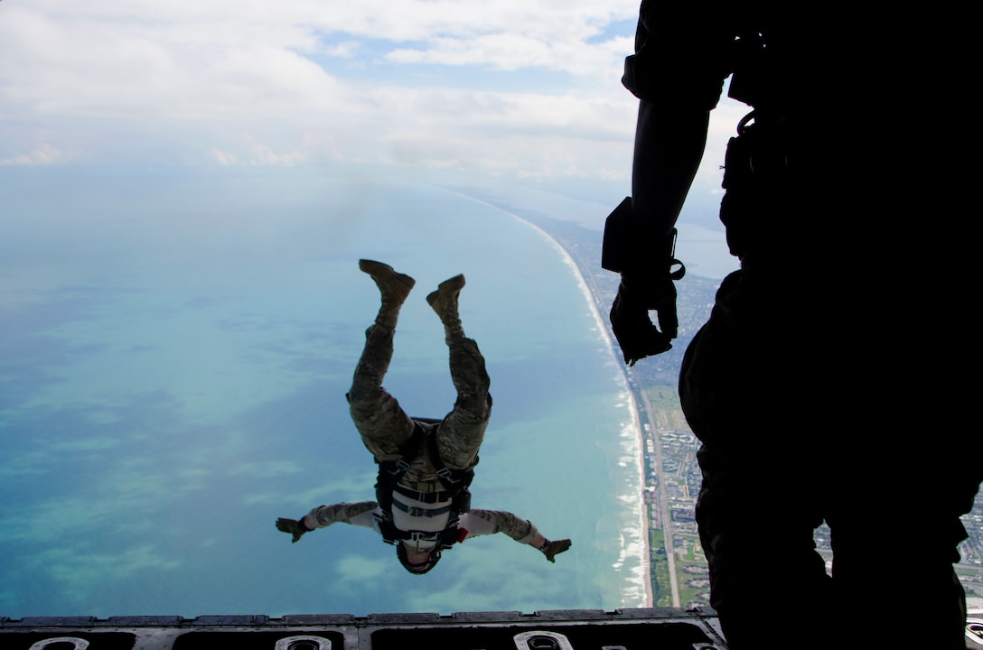 An airman falls upside-down over water as another watches from an open aircraft.