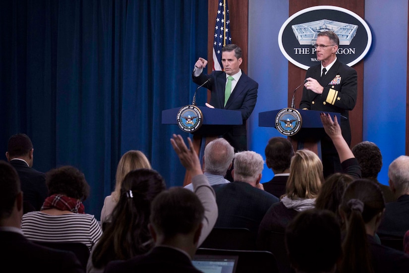 Two men stand behind lecterns. On the wall behind them, a sign reads “the Pentagon.”