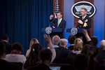 Two men stand behind lecterns. On the wall behind them, a sign reads “the Pentagon.”