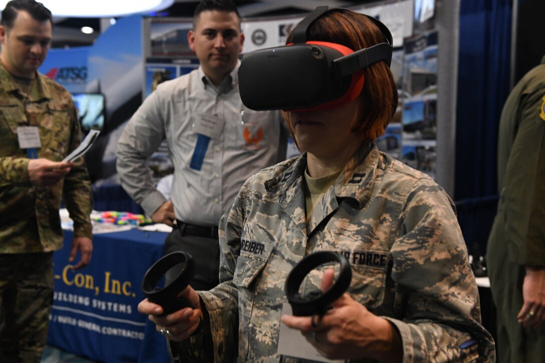 A female airman wearing virtual reality goggles holds two different handpieces. Two men at a booth in the background watch her.