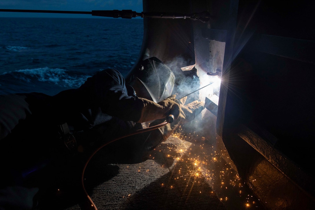A sailor lies on a ship's deck to weld something through a panel at night.