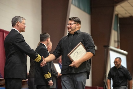 Capt. Robert Figgs, left, commanding officer, Trident Refit Facility, congratulates Shop 06 electronic industrial control mechanic Ian Jones during 2019 PSNS & IMF Apprentice Graduation ceremony Friday, Nov. 8 at the Kitsap County Fairgrounds Kitsap Sun Pavilion.