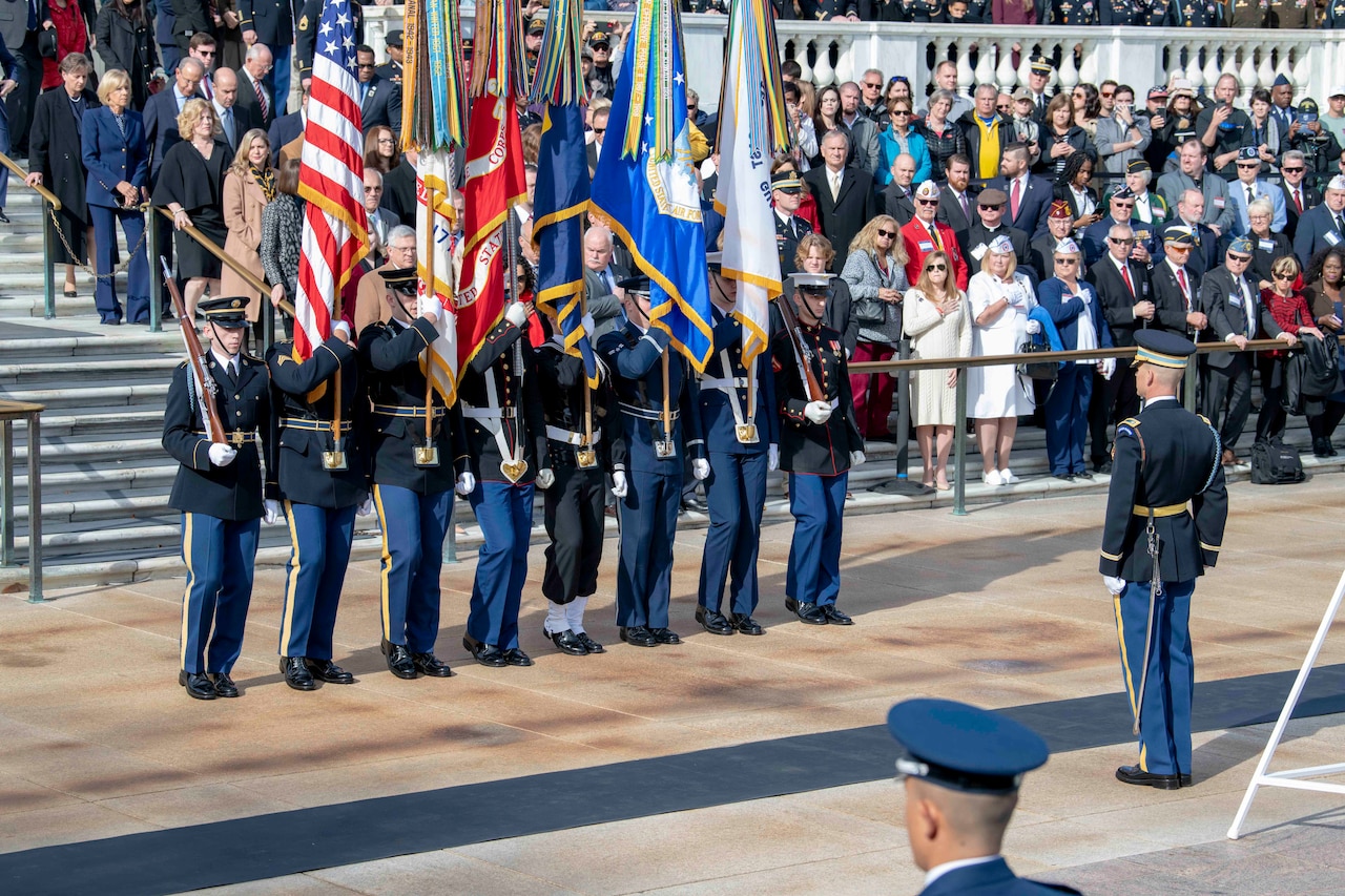 A group of service members stand in a line in front of a crowd, holding flags.