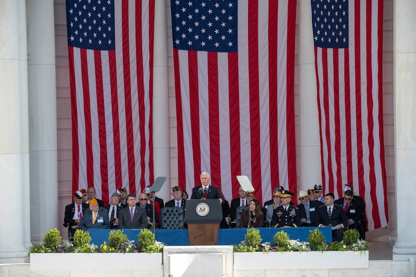 Vice President Mike Pence stands on stage with a group of people and three large American flags behind.