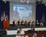 woman facilitates panel discussion of four men and three women.