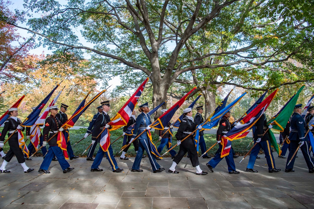 Members of all services walk in a line on an outdoor walkway with flags of different states pointed forward.