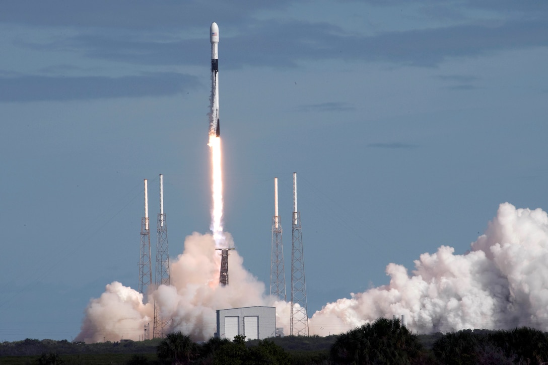 Smoke plumes spread across the ground as a rocket lifts off against blue sky.