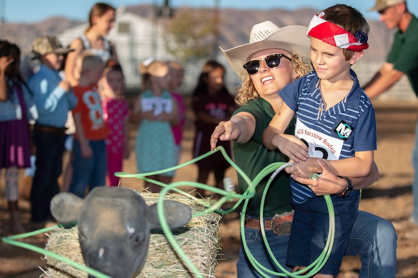 A woman wearing a white cowboy hat, shows a child wearing a red bandana how to rope a mock calf.