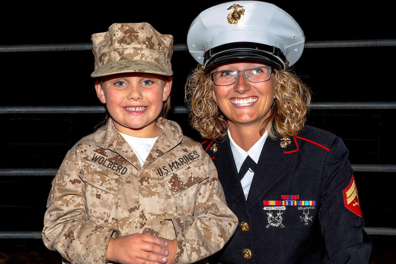 A Marine in a formal dress uniform smiles at the camera as she kneels down and has her arm around a young girl who is dressed up like a Marine.
