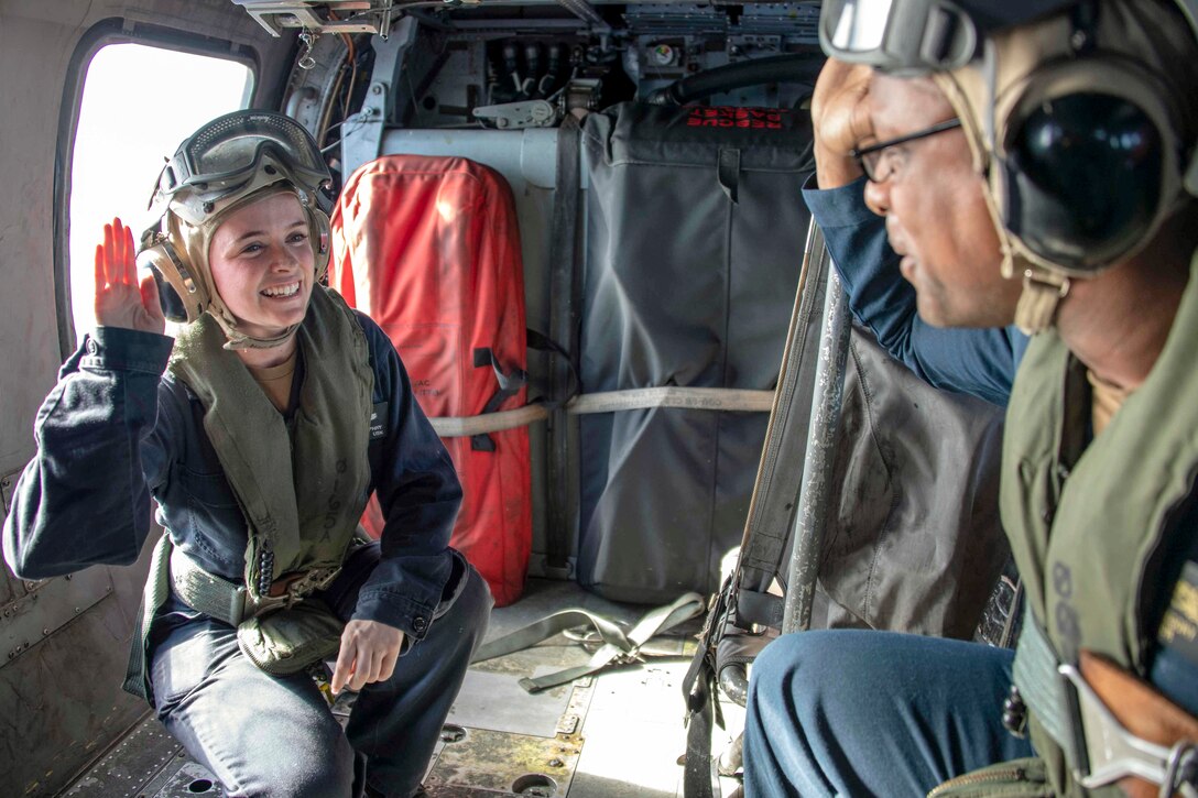 Two sailors in flight helmets raise their right hands and face each other inside a helicopter.