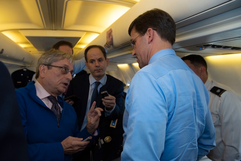 Men holding tape recorders talk to another man on board an airplane.