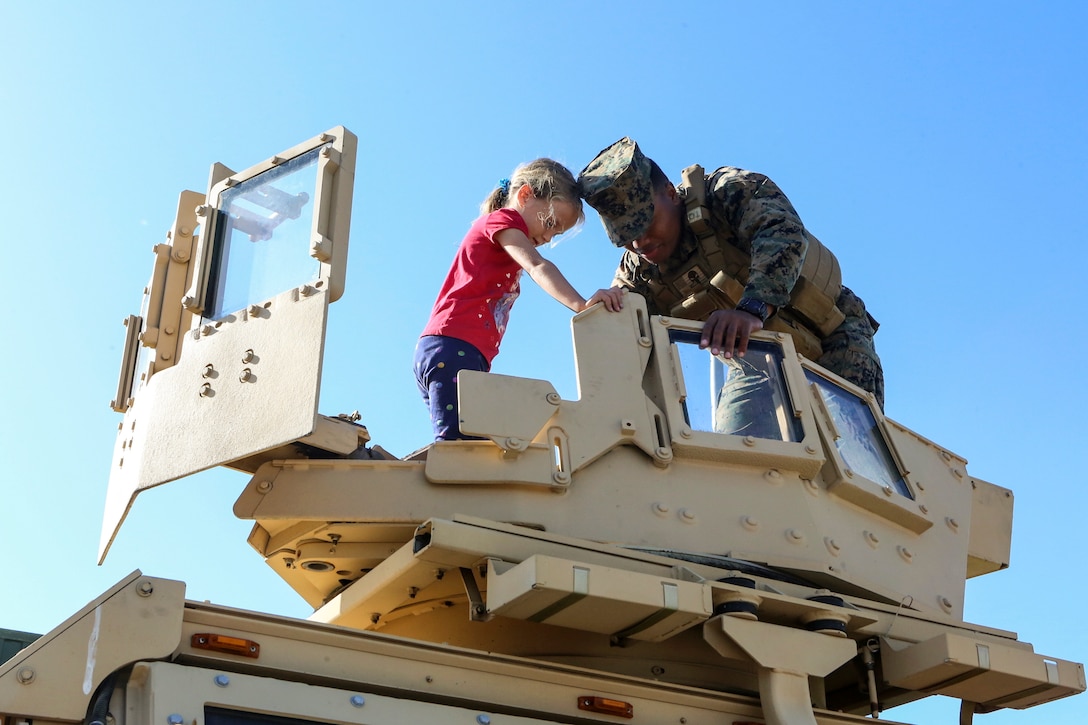 A Marine and a child stand atop a military vehicle and peer inside.