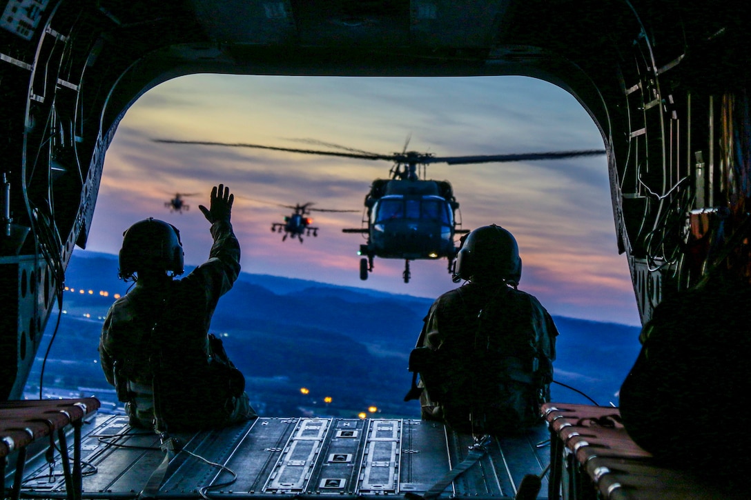 Two soldiers sit inside an aircraft as one waves at other aircraft flying behind.