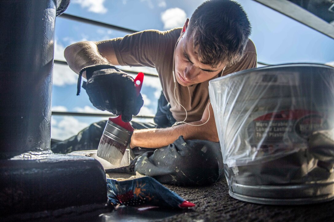 A sailor sits and paints on a ship's deck.