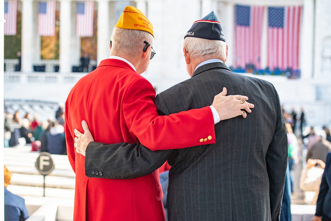 Two veterans, shown from behind, stand with their arms around each other at an amphitheater.