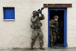 A soldier stands outside a building aiming a weapon, while another soldier holds a weapon and crouches in the doorway.