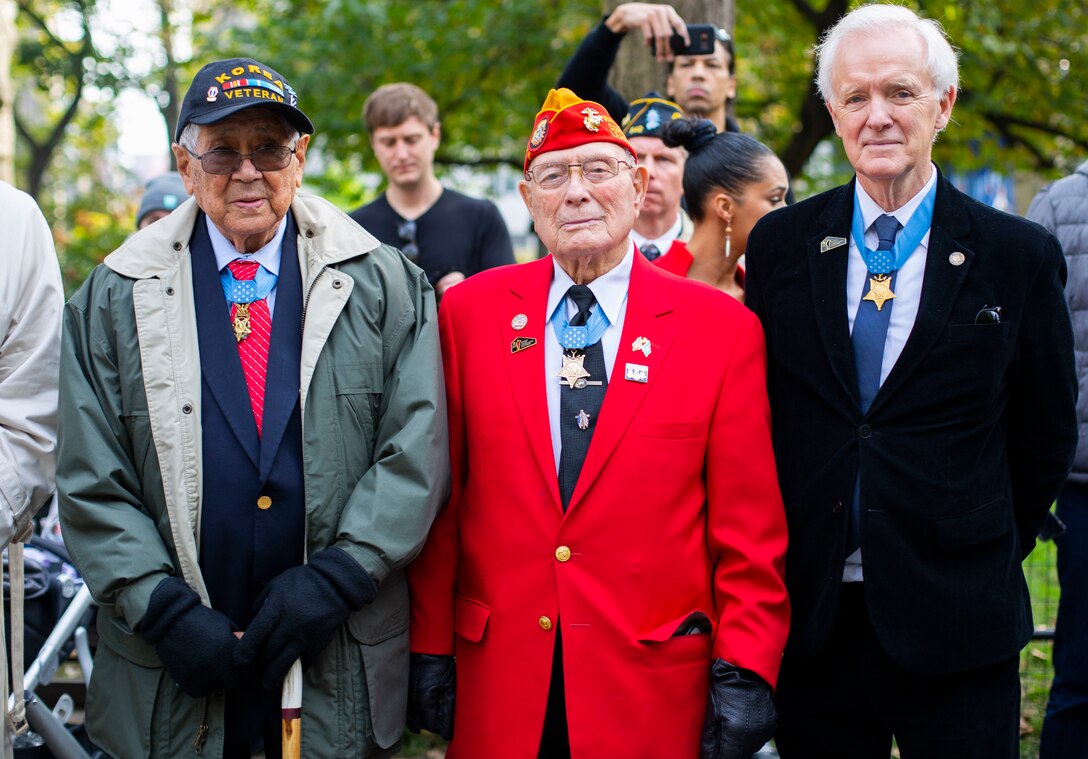 Three people wearing suits, coats and medals stand for a photo outdoors.