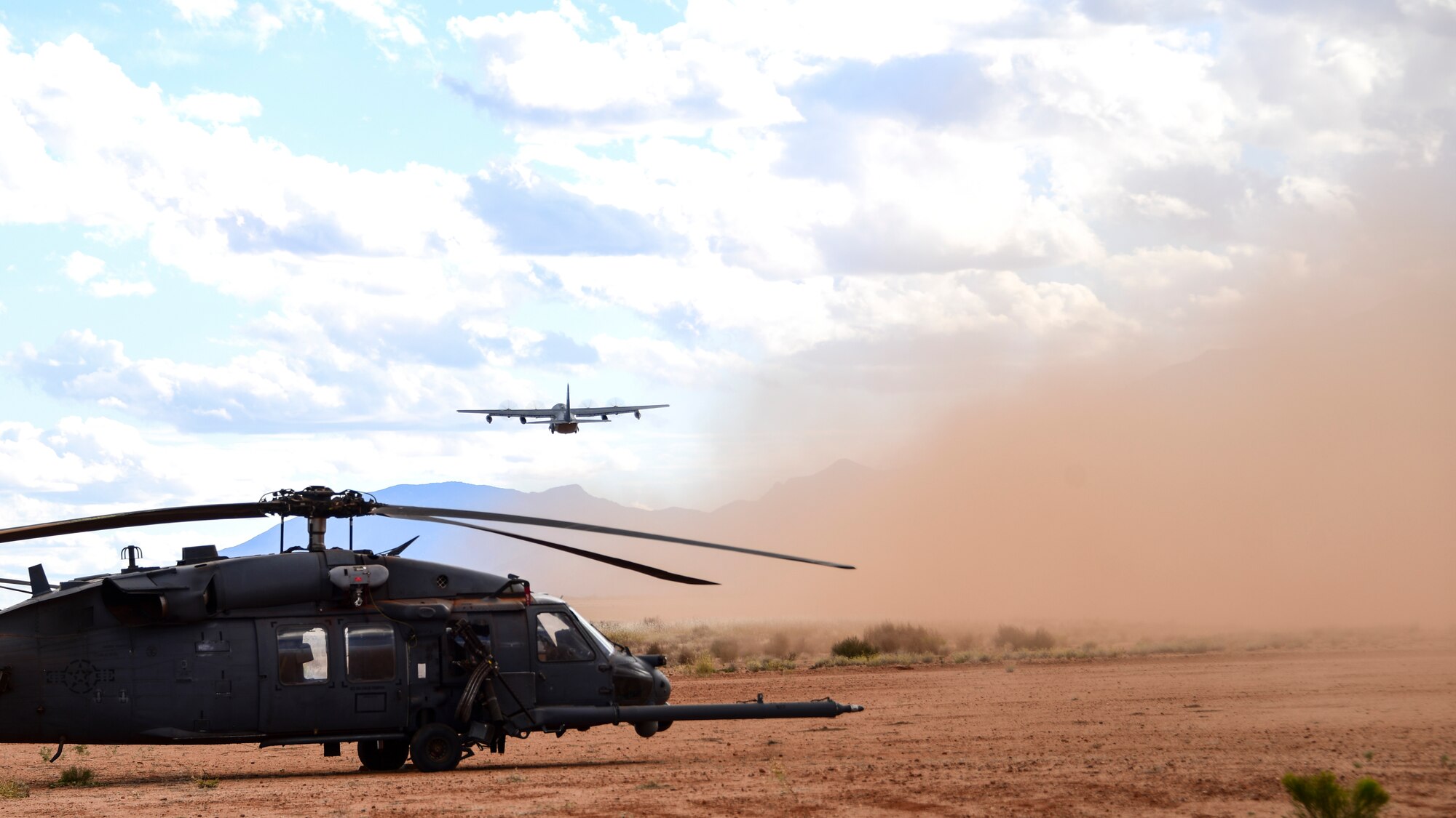 A photo of an HC-130J flying while an HH-60 sits on a dirt runway.
