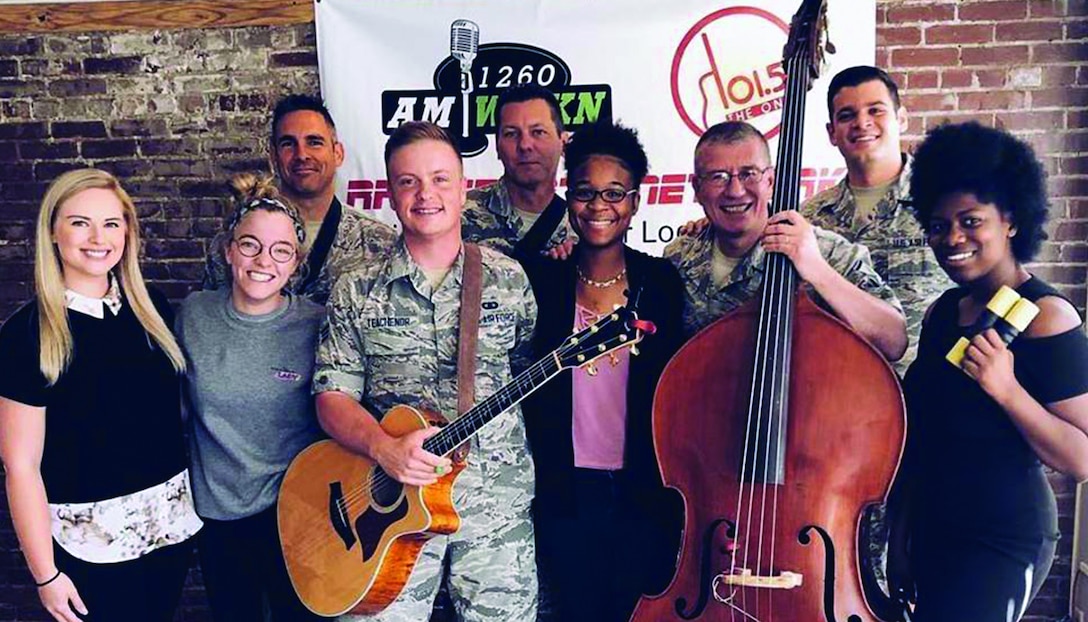Five musicians from Wild Blue Country pose with their instruments, including an upright bass and electric guitar, in their digital camoflage pattern uniforms with local musicians and engineers.