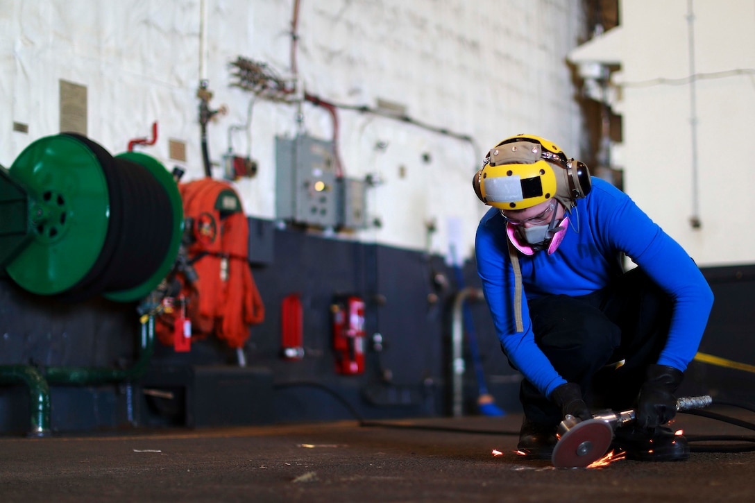 A sailor kneels on the ground using a saw on a pad eye.