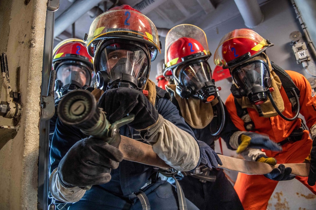 Sailors wearing fire protection gear point a hose at a simulated fire.