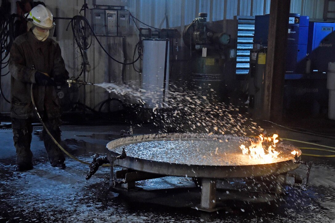 A person wearing fire protection gear sprays foam on a fire in a 28-square-foot container.