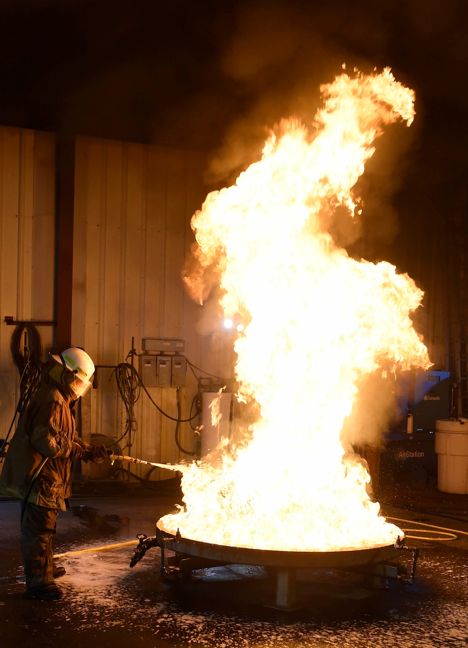 A person wearing fire protection gear sprays foam on a fire in a 28-square-foot container.