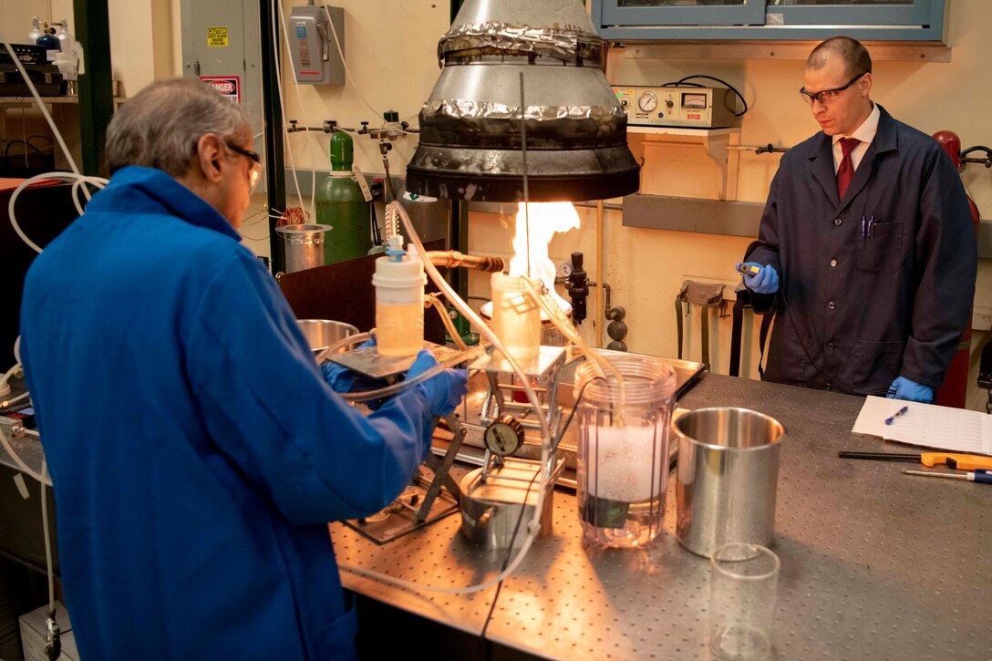 A chemist pours foam on a small fire as another chemist holds a stopwatch.