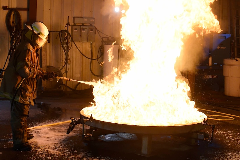 A person wearing fire protection gear sprays foam on a fire in a 28-square-foot container.