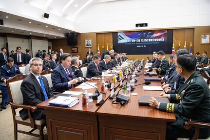 A room full of men in suits and uniform, many sitting at a large table with microphones.