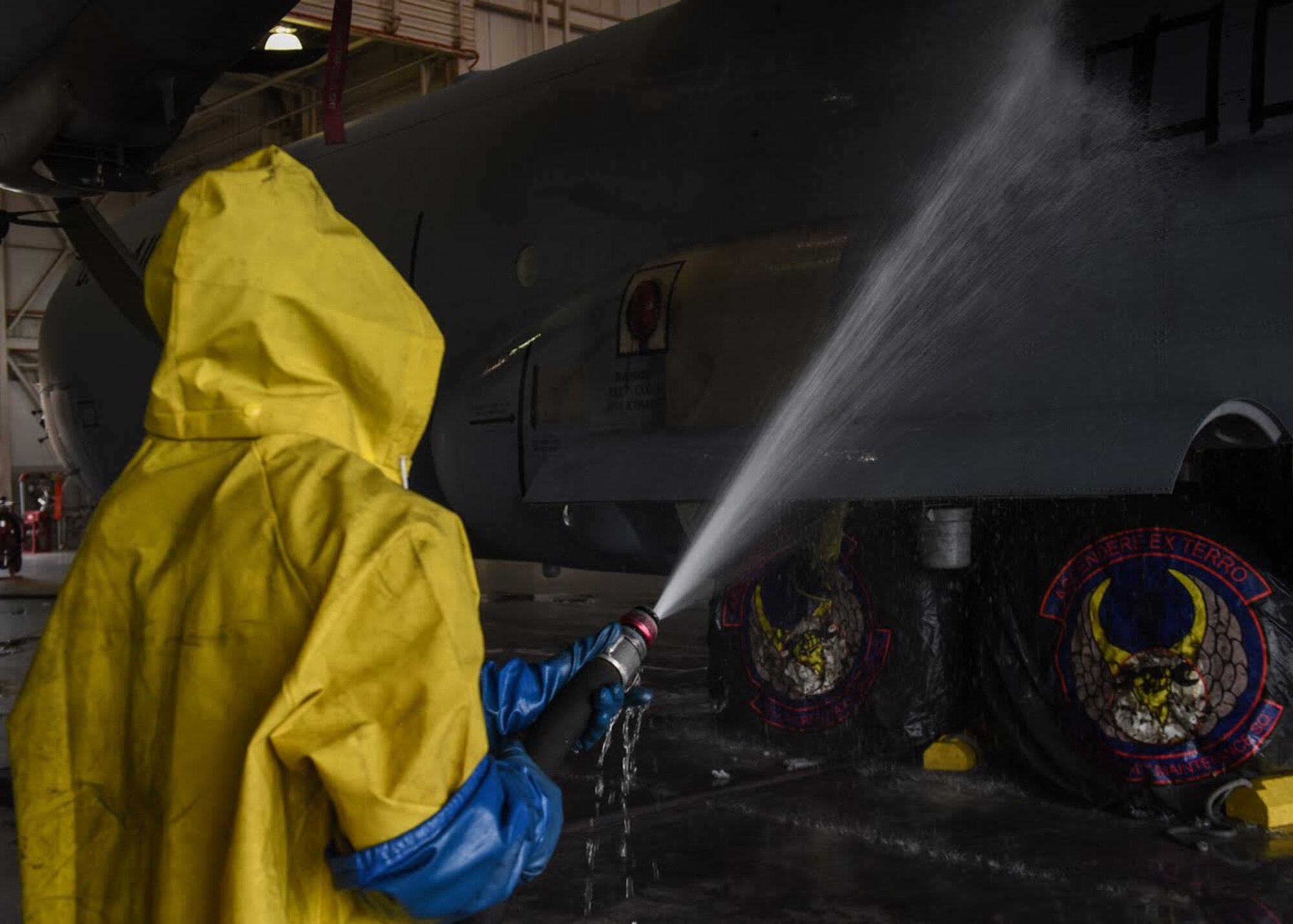 Photo shows an Airman, dressed in yellow protection gear, spraying down cleaning chemicals on the side of a C-130J Super Hercules aircraft.