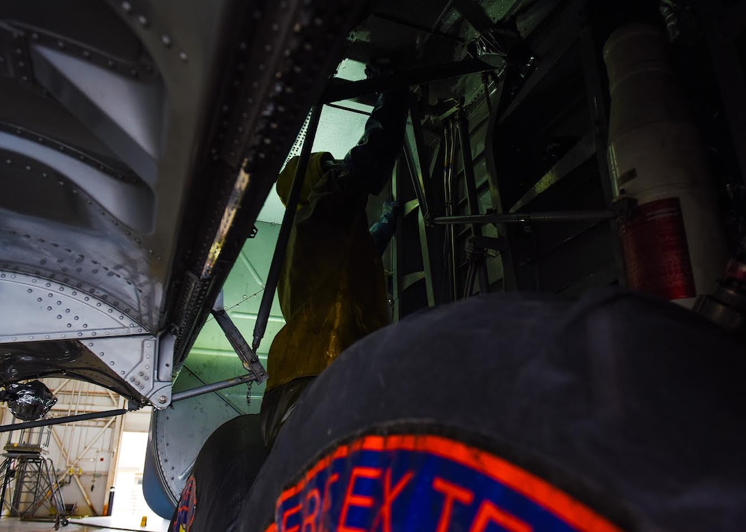 Photo shows an Airman, dressed in yellow protective gear, cleaning above the tires of a C-130J Super Hercules aircraft.