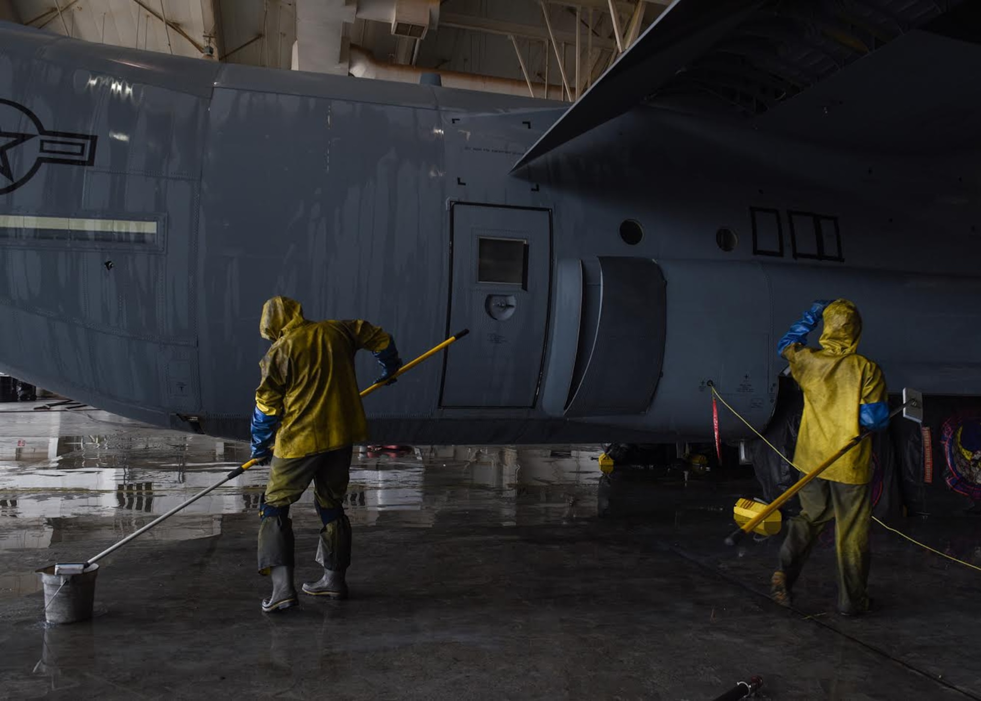 Photo shows Airmen, dressed in yellow protective gear, wetting sponges to clean a C-130J Super Hercules aircraft.